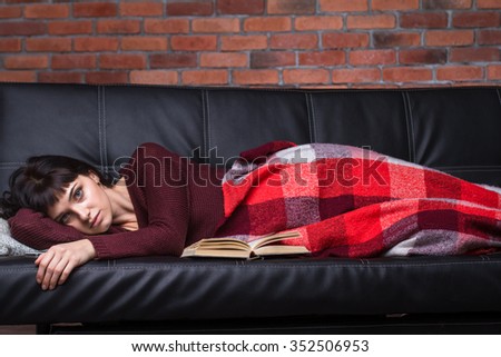 Pensive tired pretty young female lying on sofa covered with plaid coverlet over brick wall background