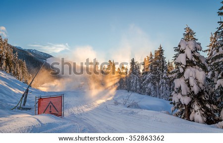 Snow-maker spraying snow onto a cat-track with sunlight coming through snowy trees. 