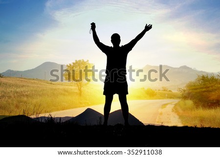 Silhouette of man tourist with arms raised with a camera on a mountain slope on the background of the road, mountains, sea and sun (double exposure)