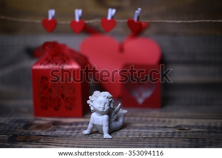 Closeup view of one beautiful cupid angel decorative figurine near red paper greeting valentine box near clothes-peg in shape of heart with no people on wooden background, horizontal picture