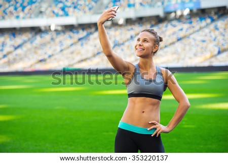 Young beautiful mixed race sportswoman making selfie photo on mobile phone after training outdoors and smiling. Fit woman is at large nice modern stadium