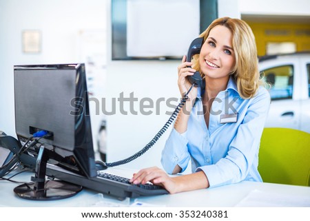 Photo of young beautiful business woman in car showroom. Young business woman working with computer and talking on phone. Modern office interior