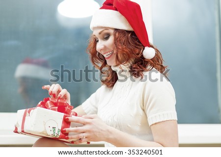 Christmas and New Year Celebration and Holiday Concept. Caucasian Red-Haired Santa Girl Unwrapping Gift Box with Positive Facial Expression. Indoors Shot. Horizontal Composition