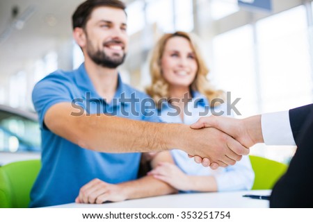 Photo of young male consultant and couple. Young woman and man buying new car in auto show. Signing of contract and shaking hands. Concept for car rental