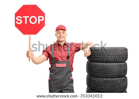 Young male mechanic holding a big red stop sign and leaning on a stack of tires isolated on white background