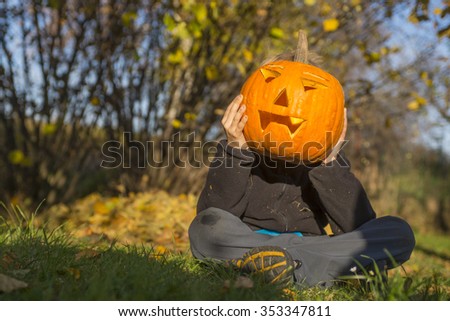 Portrait of child Boy with halloween pumpkin jack-o-lanterns in front of face, sitting on the grass, autumn garden, sunny day