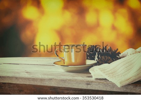 front image of coffee cup over wooden table and woolen sweater in front of autumnal sunset background. retro filtered