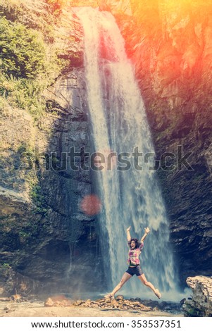 Young woman with hands up jumping near waterfall
