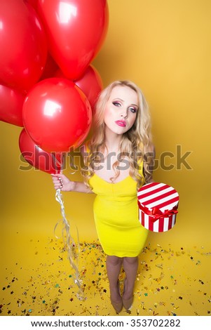 One attractive surprised young smiling blonde woman with long curly hair holding birthday round present box near bunch of red balloons with confetti in studio on yellow backdrop, vertical picture