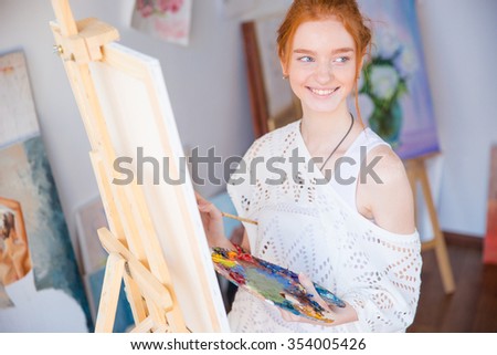 Happy lovely cute young woman painter holding palette with oil paints and painting on canvas in art studio