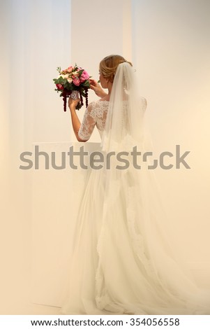 Photo of young bride full length back view wearing long lace white wedding dress and bridal veil holding beautiful decorated bouquet for ceremony in hands on pastel background, vertical picture