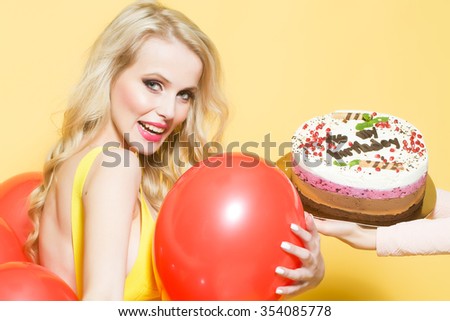 One attractive smiling young happy blond woman with long curly hair with birthday cake with candle in female hand near bunch of red party balloons in studio on yellow backdrop, horizontal picture