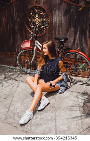 young girl in a blue t-shirt and denim shorts lips with red lipstick long blonde hair posing with red bicycle with a basket of classic vintage retro style  hipster white sneakers and a blue cape