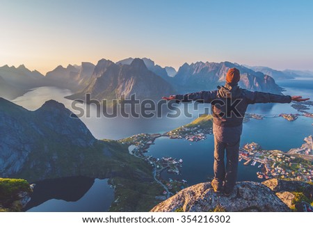 Silhouette of the man spreading arms and standing high on rock like the Statue of Christ the Redeemer looking at breathtaking view over small village,mountain landscape, sea during sunset in Norway  Royalty-Free Stock Photo #354216302