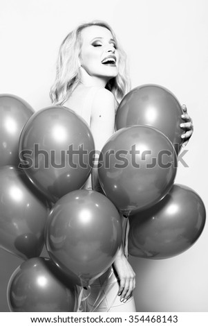 One beautiful smiling flirtatious young happy blond woman with long curly hair standing in bunch of many party balloons in studio black and white, vertical picture