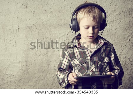 Child blond Boy listening to music or watching movie with headphones and using digital tablet. Concrete wall background