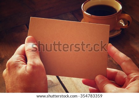 male hands holding brown empty card over wooden table background and cup of coffee. retro style image, low key and warm tones
