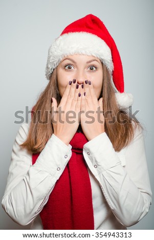 beautiful santa girl covers her mouth with her hands. Christmas hat isolated portrait of a woman on a gray background, studio photo.
