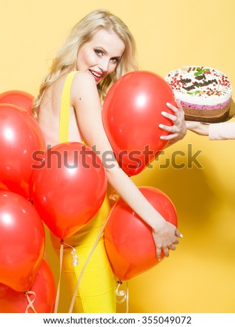 One attractive smiling young happy blond woman with long curly hair with birthday cake with candle in female hand near bunch of red party balloons in studio on yellow backdrop, vertical picture
