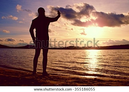Young man holding cellphone, taking picture of autumn sunset or sunrise in picturesque sea scenery