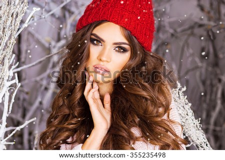Close up portrait of  beauty  winter girl in frosty winter park .Flying Snowflakes.  Beautiful young woman in red knitted hat, wavy  amazing hairstyle , full lips and  bright make up.