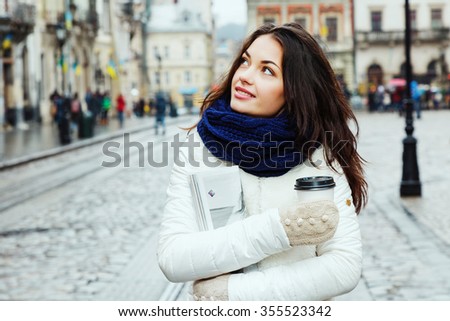 Beautiful girl, with curly long hair, wearing in white coat, blue scarf and gloves, holding cup of coffee and magazine and looking up, on the street of old European city, waist up