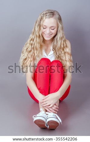 Portrait of beautiful caucasian blonde woman with curly hair standing on grey background. Girl with beautiful long hair. Fashionable girl. Red pants. Girl sitting hugging legs. Embarrassed and shy
