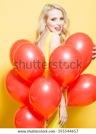 One beautiful smiling flirtatious young happy blond woman with long curly hair standing in bunch of red party balloons in studio on yellow backdrop, vertical picture