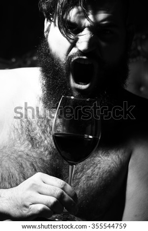 Closeup view portrait of one handsome screaming emotional young adult man with long black lush beautiful beard and moustache holding glass with red wine indoor on blurred background, vertical picture