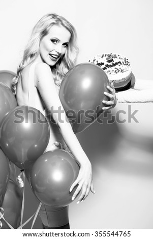 One attractive smiling young happy blond woman with long curly hair with birthday cake with candle in female hand near bunch of party balloons in studio black and white, vertical picture