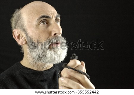 An old man, praying God with a rosary in his wrinkled hands, his head and soul looking upwards to Heaven. Black background.
