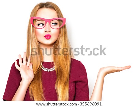 Beauty funny teenage girl with paper glasses on stick showing empty copy space on the open hand palm for text, white background. Happy girl presenting point. Proposing product. Advertisement gesture