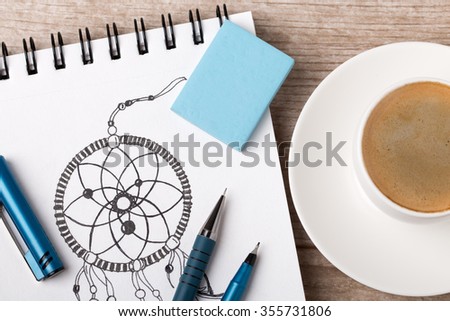 Close-up view of artist's or designer's table. Cup of coffee, pencil, fine liner and eraser laying on sketch book with hand-drawn dream catcher