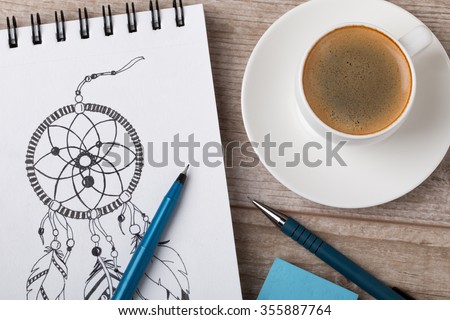 Close-up view of artist's or designer's table. Cup of coffee, pencil, fine liner and eraser laying on sketch book with hand-drawn dream catcher
