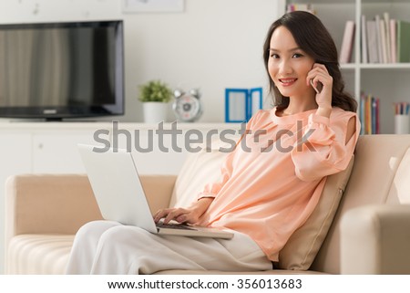 Attractive Asian woman sitting on the sofa with laptop and calling on phone