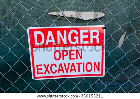 Sign says danger open excavation in red letters on a white background, hung on a fence in front of a construction site.