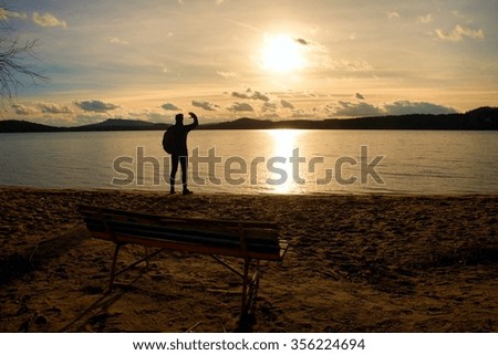 Hiker man in dark sportswear and with sporty backpack standing on the beach, relaxing and enjoy sunset at horizon with blue sky with clouds. Magic autumn day.