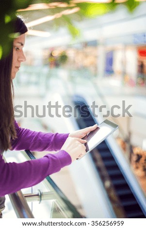  A young woman is holding a digital tablet. The image is taken in shopping center.Woman has long brown hair. Selective focus