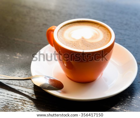Top view of a cappuccino cup with foam in the form of heart shape, saucer and silver spoon on wooden table background in the natural light of afternoon. Art coffee.