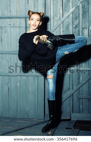 Full length view of one straight slender funny young smiling happy woman with cool hairstyle in torn jeans and black sweater with flexible body standing in studio on wooden backdrop, vertical picture