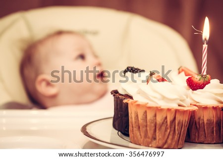 Cupcake with burning candle and sparks for first birthday and baby on background. Baby is not in focus, main focus is on cakes