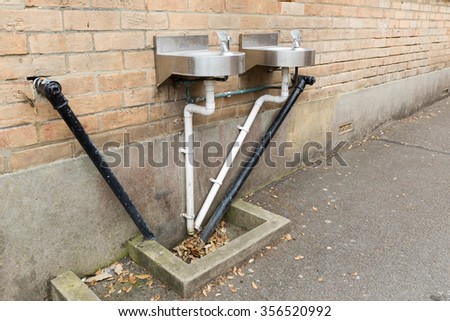 A photo of black and white angular water pipes connected to two stainless steel basins on a beige brickwork wall