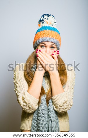 Happy beautiful girl covers  mouth with hand, knit cap, winter concept, photo studio, portrait of a woman isolated on gray background