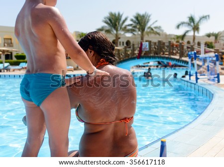 Small boy applying sunscreen on his mother rubbing it into her back and shoulders as she relaxes at the edge of a resort swimming pool on summer vacation