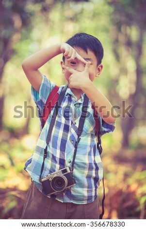 Asian boy with professional digital camera on blurred nature background. Child making frame with hands, taking picture with imaginary camera, selective focus. Photo in retro style. Outdoors portrait.