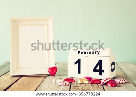 February 14th wooden vintage calendar with colorful heart shape chocolates next to blank vintage frame on wooden table. selective focus.Template ready to put photography. retro filtered
