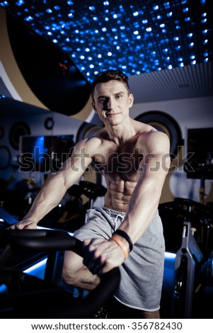 Beautiful sport bodybuilder man on a diet, cardio training on the simulator led. With six pack, perfect brakes, shoulders, biceps, triceps and chest. Young adult man doing exercises in the gym.
