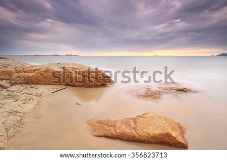 Long exposure of wave hitting rock during twilight with stunning sky and clouds. Visible in distance heavy industry pier and crane. Nature composition. Image taken at Teluk Batik, Perak, Malaysia.