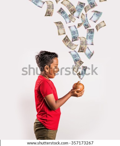 10 year old indian boy excited to see currency notes falling in clay money box, asian boy holding piggy bank and money falling into it, indian currency flying, indian rupee falling Royalty-Free Stock Photo #357173267