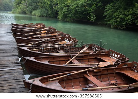 Photo closeup of row of identical wet wooden brown boats with oars mooring with chains at timber pier on beautiful riverscape background, horizontal picture 